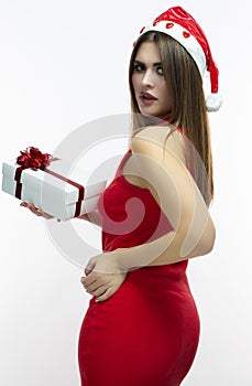 Christmas Ideas. Caucasian Girl In Red Dress and Santa Hat. Holding White Gift Box Wrapped Up with red Ribbon.