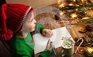 Christmas Kid writing Letter to Santa. Happy Child in Red Santa Hat dreaming about Presents at Home. Xmas Gifts Shopping Wish List