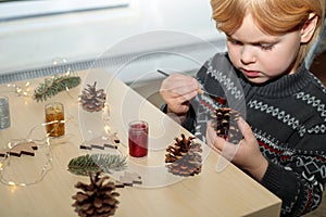 Christmas kids craft activities at home. Little child painting pine cones and wooden Christmas tree decorations