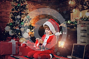 Christmas Kids. Happy little kid is wearing Santa clothes, playing with Christmas gift box. Fireplace background