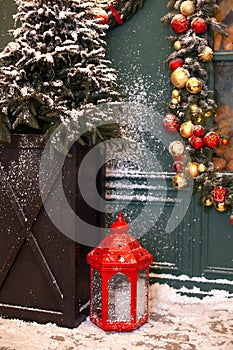 Christmas lantern in snow with a Christmas tree and garland with toys in evening scene. Christmas lantern stands in backyard on te