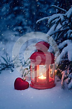 Christmas lantern under the tree with a gift in the woods. Christmas eve in the woods. Christmas lantern and toy heart in the snow