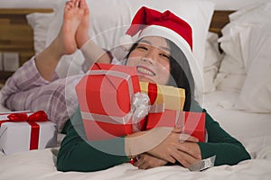 Christmas lifestyle portrait of young beautiful and happy Asian Chinese woman on bed in Santa Claus hat holding lot of xmas
