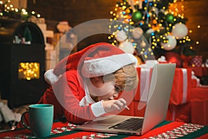 Christmas little boy typing letter to Santa Claus on a computer at home on Christmas background. Boy wearing Santa