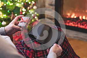 Christmas. Man using tablet for searching gift ideas sitting with ready gift boxe near fireplace and christmas tree. Concept.