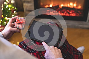 Christmas. Man using tablet for searching gift ideas sitting with ready gift boxe near fireplace and christmas tree. Concept