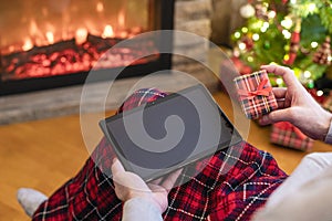 Christmas. Man using tablet for searching gift ideas sitting with ready gift boxe near fireplace and christmas tree. Concept.