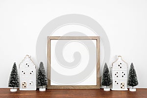 Christmas mock up with wooden frame, trees and white house decor on a wood shelf against a white wall