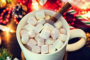 Christmas mood - white mug of cocoa with marshmallows and cinnamon stick close-up, Christmas tree branch with garland