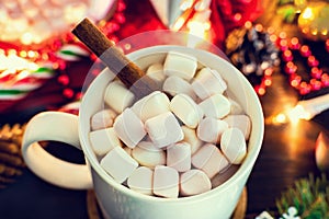 Christmas mood - white mug of cocoa with marshmallows and cinnamon stick close-up, Christmas tree branch with garland