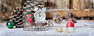 Christmas and New year alarm clock with snow on wooden sled with cones and toys, banner