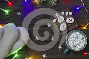 Christmas New Year card. Cup of hot drink with marshmallows, warm white mittens, snowmen on a dark wooden background. Festive