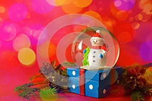 Christmas and new year card with snow globe snowman inside. Gift box on red bokeh background. Holidays, winter and celebration con