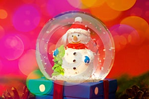 Christmas and new year card with snow globe snowman inside. Gift box on red bokeh background. Holidays, winter and celebration con