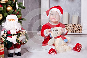 Christmas or New year celebration. Little girl in red dress and santa hat with bear toy sitting on the floor near the Christmas tr