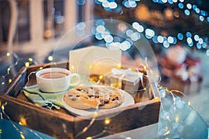 Christmas and New Year composition. Cocoa, cookies, little giftbox and postcard on the wooden tray with xmas tree background.