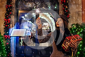 Christmas, New Year concept. Woman burning sparkler on city street by decorations. Girl holding gift box under snow.