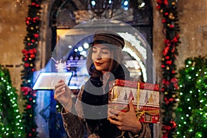 Christmas, New Year concept. Woman burning sparkler on city street by decorations. Girl holding gift box under snow