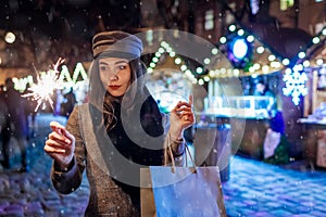 Christmas, New Year concept. Woman burning sparkler on city street fair. Girl holding paper bags with presents.