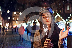 Christmas, New Year concept. Woman burning sparkler on city street fair. Girl holding paper bags with presents