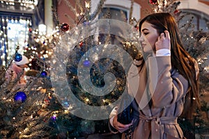Christmas, New Year concept. Woman walking on city street by decorated trees. Stylish girl enjoying holiday atmosphere