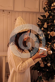 Christmas, New Year. Pretty young woman in white sweater and knitted hat  with  luminous gift box near christmas tree,  new year m