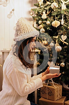 Christmas, New Year. Pretty young woman in white sweater and knitted hat  with  luminous gift box near christmas tree,  new year m