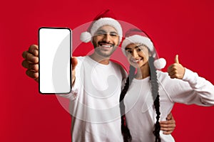 Christmas Offer. Happy Arab Couple Wearing Santa Hats Demonstrating Blank Smartphone Screen