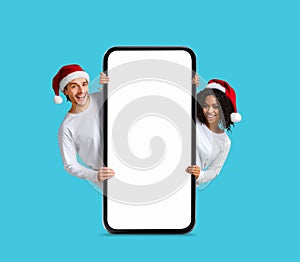 Christmas Offer. Happy Multiethnic Couple In Santa Hats Standing Behind Blank Smartphone