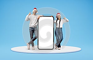 Christmas Offer. Indian Couple Wearing Santa Hats Standing Near Big Blank Smartphone