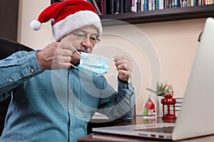 Christmas online congratulation. Senior man in santa claus hat put on face mask and talks using laptop device. The room is