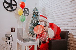 Christmas online congratulation. Young man in Santa Claus costume and face mask gives a gift and talks using laptop for video call