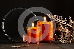 Christmas orange burning candles in orange glass in darkness with decoration of dry leaves and mirror on black wood table
