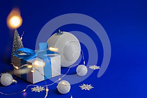 Christmas ornaments. White gift box with blue ribbon, winter tree, Snowflakes and Silver balls in xmas decorations on blue