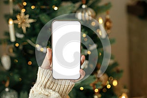 Christmas phone mock up. Hand holding smartphone with empty screen against stylish festive christmas tree with golden lights.