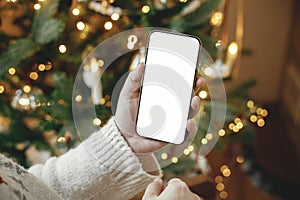 Christmas phone mock up. Hand holding smartphone with empty screen against stylish festive christmas tree with golden lights.