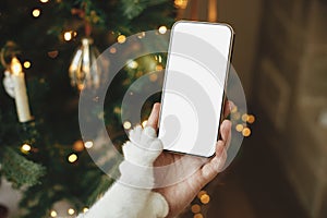 Christmas phone mock up. Hand holding smartphone with empty screen and cute cat paw against stylish christmas tree with golden
