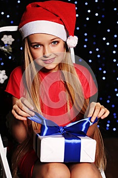Christmas photo of cute little blond girl in santa hat and red dress holding a gift - box on the backgroud of holiday shining ligh