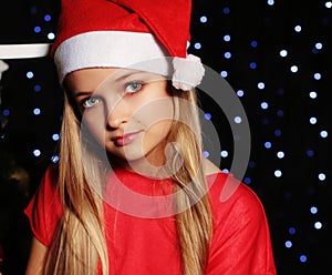 Christmas photo of cute little blond girl in santa hat and red dress holding a gift - box on the backgroud of holiday shining ligh