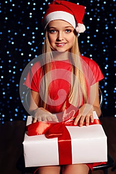 Christmas photo of cute little blond girl in santa hat and red dress holding a gift - box on the backgroud of holiday shining ligh