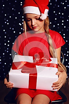 Christmas photo of cute little blond girl in Santa hat and red dress holding a gift - box