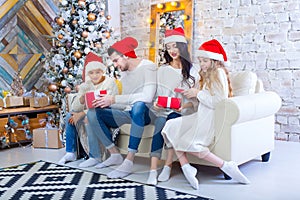 A Christmas photograph of a beautiful family with two children in Santa Claus red caps and red presents. Sit on the