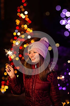 Christmas photography. the girl holds a sparkler in her hand, the lights of garlands on the tree are visible from behin