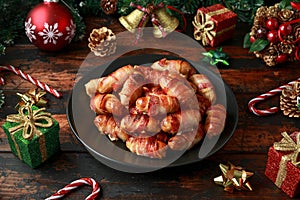 Christmas Pigs in blankets, sausages wrapped in bacon with decoration, gifts, green tree branch on wooden rustic table