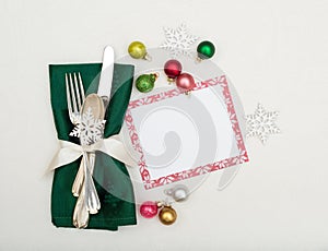 Christmas Place Setting with Silverware in Green Napkin on Off White Table Cloth Background with menu card and colorful ornaments