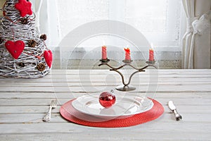 Christmas place setting with white dishware, cutlery, silverware and red decorations on white.