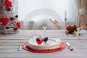 Christmas place setting with white dishware, cutlery, silverware and red decorations on white wooden board. Christmas