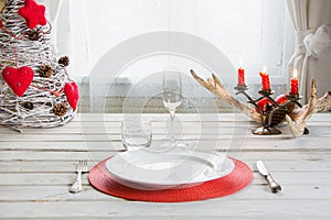 Christmas place setting with white dishware, cutlery, silverware and red decorations on white wooden board. Christmas