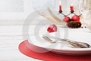 Christmas place setting with white dishware, cutlery, silverware and red decorations on wooden board. Christmas