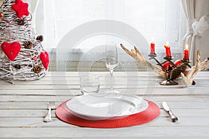 Christmas place setting with white dishware, silverware and decorations on white board in interior near the window.